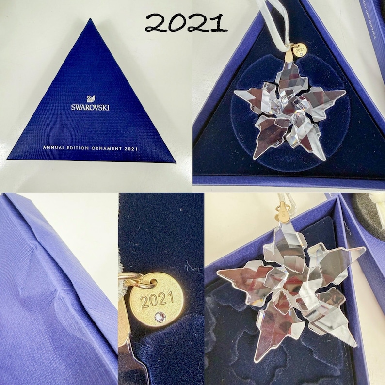 May include: A clear crystal star-shaped ornament with a white ribbon hanger and a small gold-tone metal tag engraved with 2021. The ornament is presented in a triangular blue box labeled Swarovski Annual Edition Ornament 2021.