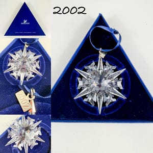 May include: A clear crystal snowflake ornament with sharp, faceted points, featuring a small metal tag engraved with the year 2002. The ornament includes a dark blue ribbon for hanging and is presented in a triangular dark blue velvet-lined box.