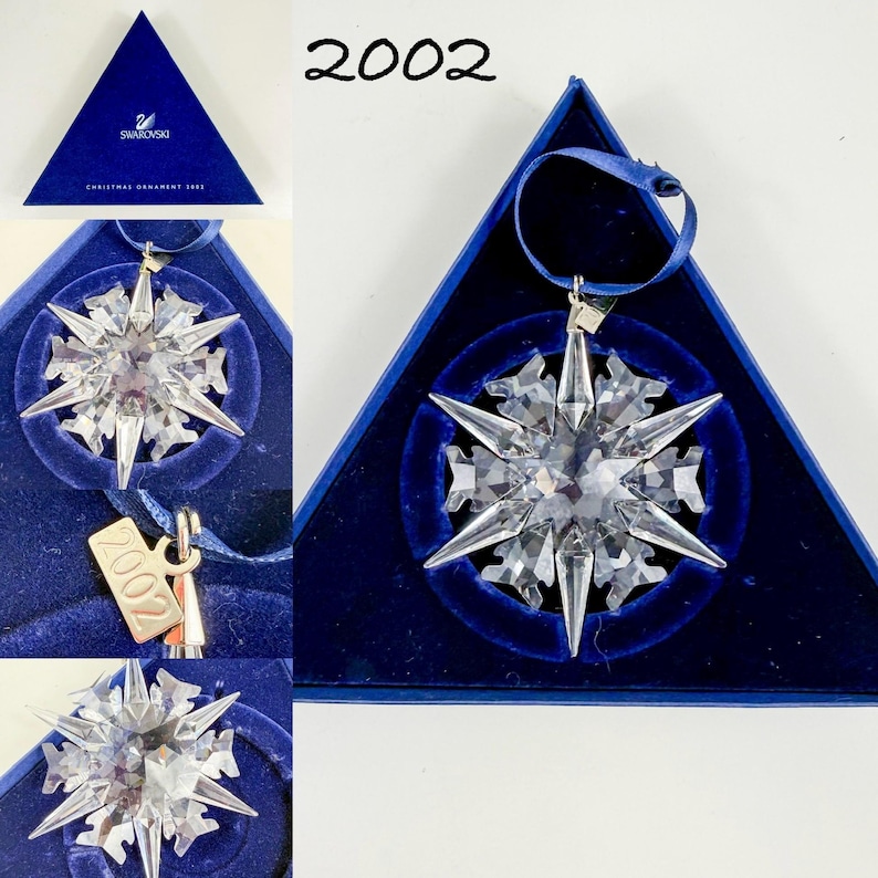 May include: A clear crystal snowflake ornament with sharp, faceted points, featuring a small metal tag engraved with the year 2002. The ornament includes a dark blue ribbon for hanging and is presented in a triangular dark blue velvet-lined box.