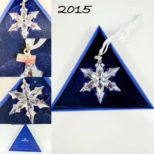May include: A clear, faceted crystal snowflake ornament with a white ribbon hanger. A small metal tag attached to the top is engraved with the year 2015. The ornament is presented in a triangular blue box with a dark blue velvet interior lining.