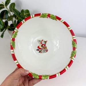 Op de afbeelding: Een witte keramische kerstkom met een rood-witte gestreepte rand en hulstbladaccenten. De kom heeft een illustratie van de kerstman in het midden. Een hand houdt de kom vast.