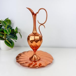 May include: A copper-colored pitcher with a curved handle and a bulbous body, set on a matching scalloped tray. The pitcher has a long, slender spout. The items are displayed on a white surface, with a green plant in a white pot in the background.