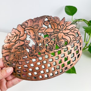 May include: An oval copper-coloured decorative piece with a floral design. It features roses, lotus flowers, and other floral elements, plus a lattice-style base. The metal has a slightly aged appearance.