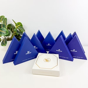 May include: A collection of triangular blue Swarovski gift boxes with the brand logo and text, arranged behind a single white octagonal box featuring a gold-colored swan logo. The blue boxes are labeled with various years including 2021, 2020, and 2018.