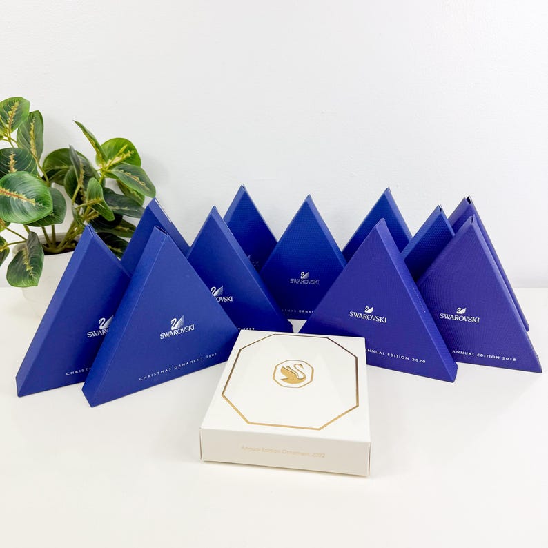 May include: A collection of triangular blue Swarovski gift boxes with the brand logo and text, arranged behind a single white octagonal box featuring a gold-colored swan logo. The blue boxes are labeled with various years including 2021, 2020, and 2018.