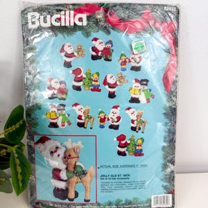 Könnte beinhalten: Ein Bucilla Jolly Old St. Nick Filz-Ornament-Set. Das Paket enthält ein Set mit 12 Filz-Ornamenten, die jeweils etwa 10 cm hoch sind. Die Ornamente zeigen den Weihnachtsmann, Rentiere und andere festliche Figuren.