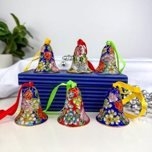 Puede incluir: Seis adornos de campana decorativos con diseños florales en rojo, azul, blanco y verde. Cada campana tiene una cinta colorida para colgar. Las campanas están dispuestas sobre una caja a rayas azules, lo que sugiere un tema festivo.