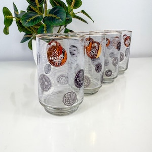 May include: Set of five vintage highball glasses. Each glass features a gold rim and is decorated with coin-themed designs in shades of brown, gold, and purple. The glasses have a clear, textured appearance.