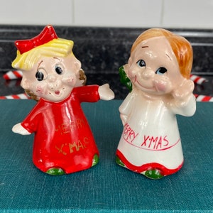 May include: Two vintage ceramic Christmas figurines. One is a girl with blonde hair wearing a red dress with the words "Merry Xmas" on it. The other is a girl with brown hair wearing a white dress with the words "Merry Xmas" on it.