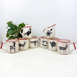 以下が含まれることがあります： クリーム色の陶器製マグカップとピッチャーのセット。それぞれ黒い動物のイラストと赤い斑点模様の縁取りが施されています。セットには七面鳥が描かれた蓋付き容器、亀が描かれたピッチャー、雄羊、鹿、鷲、ウサギ、鳥が描かれたマグカップが含まれています。