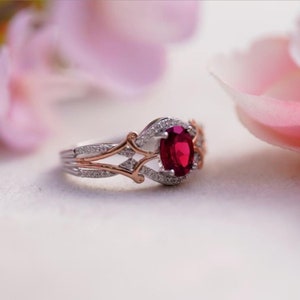 May include: A close-up of a ring featuring a large, oval-cut ruby gemstone. The ring has a silver band with rose gold accents and is encrusted with small, sparkling diamonds. The design is intricate and elegant, showcasing a blend of classic and modern styles.