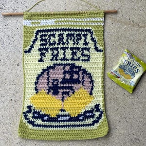 Puede incluir: Tapiz de pared tejido a ganchillo con forma de bolsa de Scampi Fries. El diseño verde, amarillo y blanco presenta las palabras "SCAMPI FRIES" en azul marino. Incluye una varilla de madera y una cuerda para colgar. Una bolsa de Scampi Fries está al lado.