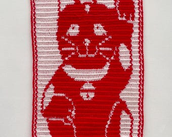 Puede incluir: Un tapiz de pared de ganchillo rojo y blanco con la imagen de un gato saludando. El gato es rojo con detalles blancos y un borde rojo. Las palabras "LUCKY CAT" están en la parte inferior. Una clavija de madera y una cuerda roja en la parte superior para colgar.