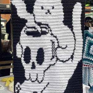 May include: A black and white crochet wall hanging featuring a cat holding a skull. The cat and skull are white, set against a black background. The handmade piece is displayed at an outdoor market.