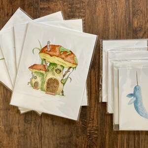 May include: Set of 5 blank greeting cards featuring watercolor illustrations. One card shows a whimsical mushroom house with a brown door and green vines. Another card shows a blue narwhal with a white horn.