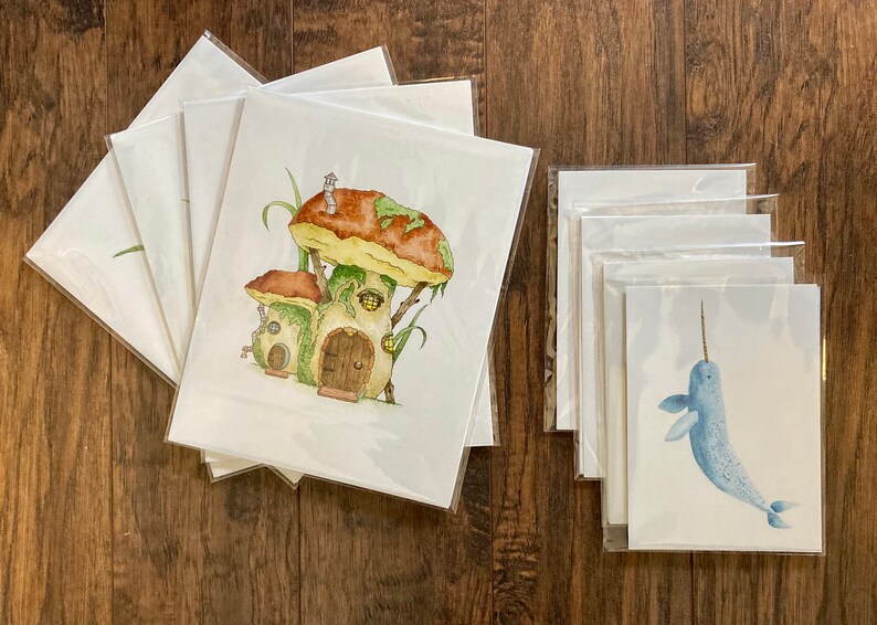 May include: Set of 5 blank greeting cards featuring watercolor illustrations. One card shows a whimsical mushroom house with a brown door and green vines. Another card shows a blue narwhal with a white horn.