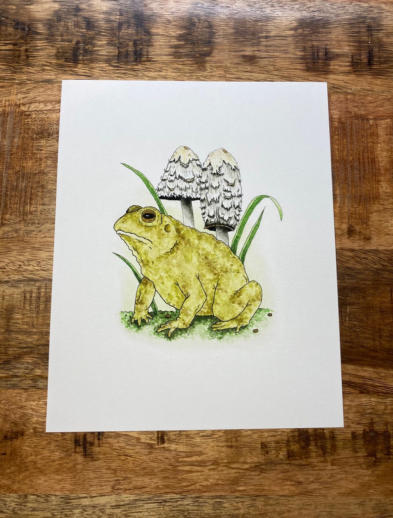 May include: A watercolor painting of a yellow toad sitting on a bed of green grass with two white mushrooms behind it.