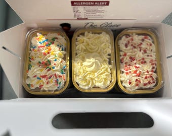 May include: A box containing three individual desserts in clear plastic containers. Each dessert is topped with a generous swirl of white frosting. One is decorated with colorful sprinkles, another with white frosting, and the third with red sprinkles.