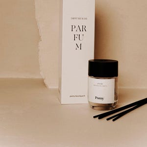 Może przedstawiać: Białe prostokątne pudełko z napisem "DIFFUSEUR DE PARFUM" i szklana butelka z czarną zakrętką. Butelka ma etykietę z napisem "AMORE" i "Parfum". Czarne patyczki umieszczone w pobliżu butelki. Tło jest w kolorze jasnobeżowym.