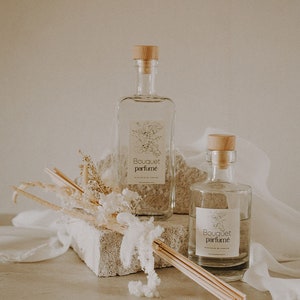 Puede incluir: Dos botellas de vidrio transparente con tapones de madera y etiquetas que dicen "Bouquet parfumé" sobre una superficie marrón claro. Las botellas están llenas de un líquido transparente y están rodeadas de flores blancas secas y un manojo de palitos de madera.