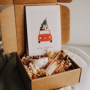 Könnte beinhalten: Ein brauner Karton mit offenem Deckel, der eine kleine weiße Kerze, ein Glasröhrchen mit Streichhölzern und eine Karte mit einem roten Auto zeigt, das einen Weihnachtsbaum und Geschenke transportiert. Die Karte lautet "Frohe Weihnachten".