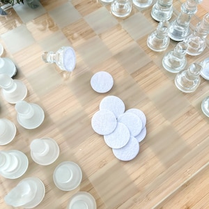 May include: A glass chess set on a wooden board with white felt pads. The chess pieces are arranged in a playing position.