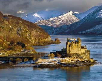 May include: A stone castle sits on a small island in a body of water with snow-capped mountains in the background. The castle is surrounded by water and a small bridge leads to the island.