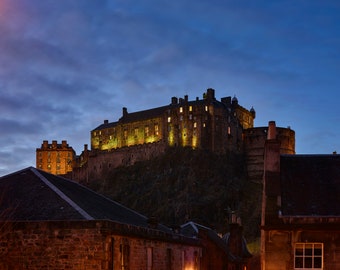 May include: A view of Edinburgh Castle at dusk, illuminated by warm lights against a dark blue sky. The castle is perched on a hilltop, with a smaller building visible in the foreground.