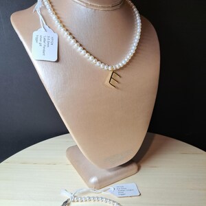 Peut inclure: Un collier de perles avec un pendentif lettre "E" doré est présenté sur un mannequin. Un bracelet de perles assorti avec un charm argenté est également montré. Les bijoux sont présentés sur une surface en bois.