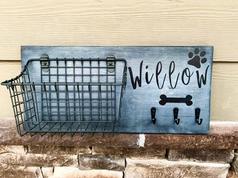 May include: A grey wooden sign with the name "Willow" and a paw print. The sign has a black metal wire basket and four hooks for leads.