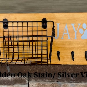 May include: A wooden dog leash holder with a wire basket, three hooks, and the name "JAY" with a paw print in silver vinyl. The wood is stained in a golden oak colour.