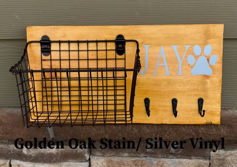 May include: A wooden dog leash holder with a wire basket, three hooks, and the name "JAY" with a paw print in silver vinyl. The wood is stained in a golden oak colour.