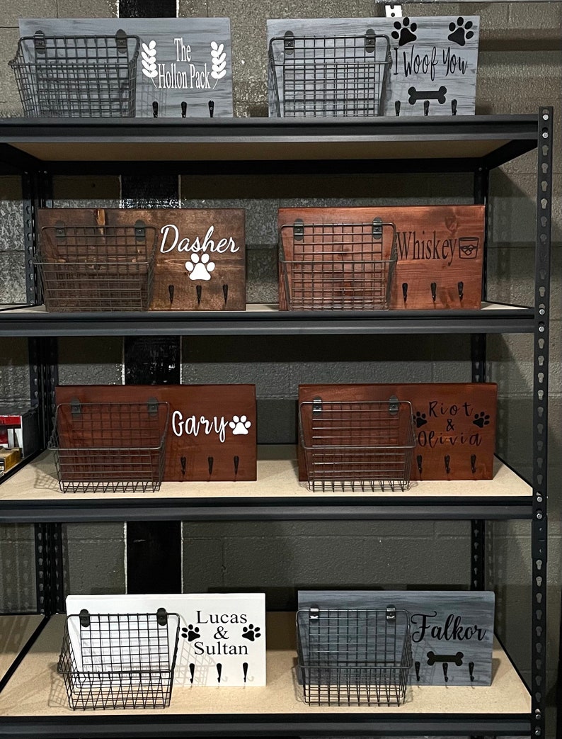 May include: A shelf unit with multiple wooden signs featuring dog names and paw prints. Each sign has a wire basket attached. The signs are in various colours, including grey, brown, and white.