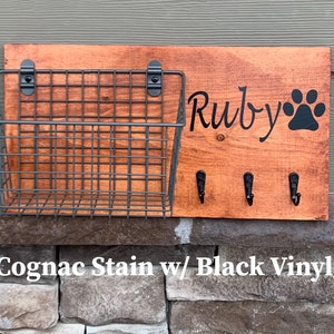 May include: A wooden dog leash holder with a wire basket, three hooks, and the name "Ruby" with a paw print. The holder is stained in a cognac colour and has black vinyl lettering.