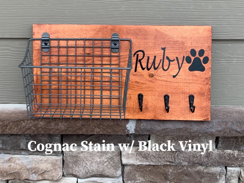 May include: A wooden dog leash holder with a wire basket, three hooks, and the name "Ruby" with a paw print. The holder is stained in a cognac colour and has black vinyl lettering.