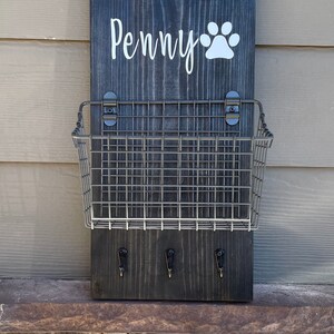 May include: A black wooden sign with the name "Penny" and a white paw print. The sign has a metal wire basket attached to it and three hooks underneath.