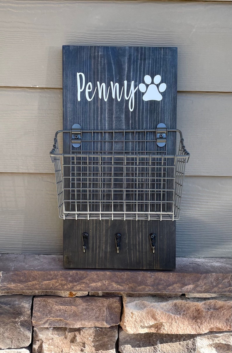May include: A black wooden sign with the name "Penny" and a white paw print. The sign has a metal wire basket attached to it and three hooks underneath.