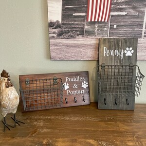 May include: Two wooden key holders with metal baskets and hooks. The key holder on the left says "Puddles & Poptart" with a paw print on each side of the text. The key holder on the right says "Penny" with a paw print to the right of the text.