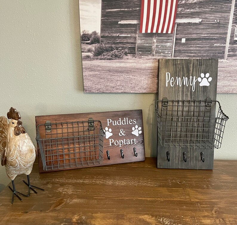 May include: Two wooden key holders with metal baskets and hooks. The key holder on the left says "Puddles & Poptart" with a paw print on each side of the text. The key holder on the right says "Penny" with a paw print to the right of the text.