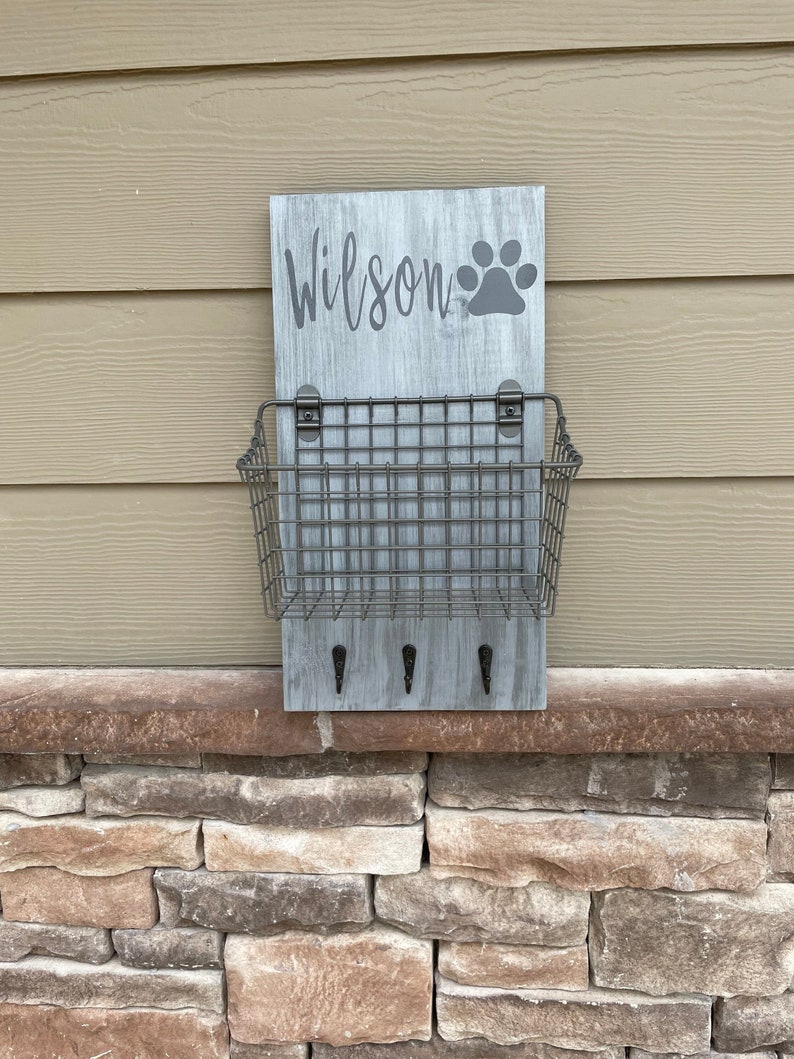 May include: A gray wooden sign with the name "Wilson" and a paw print, attached to a metal wire basket with three hooks underneath. The sign and basket are mounted on a light brown wall.