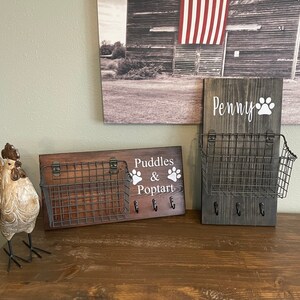 May include: Two wooden key hooks with wire baskets and paw print designs. The top hook says "Penny" and the bottom hook says "Puddles & Poptart".