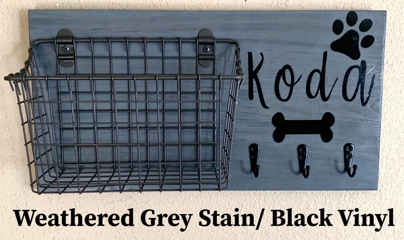 May include: A weathered grey wooden dog leash holder with a black wire basket, three hooks, and black vinyl lettering that says "Koda" with a paw print and bone design.