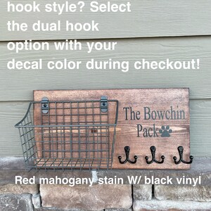 May include: A wooden sign with the text "The Bowchin Pack" and a paw print. The sign has three black hooks and a wire basket underneath. The sign is stained red mahogany with black vinyl lettering.