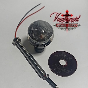 Könnte beinhalten: Ein Satz von Arcade-Stick-Komponenten, darunter ein klarer, kugelförmiger Joystick-Aufsatz mit einem Porträt, ein silberner Schaft und ein rotes und schwarzes Kabel. Eine dunkelrote Scheibe und das Logo "Vampiremul Arcade Sticks" sind ebenfalls zu sehen.