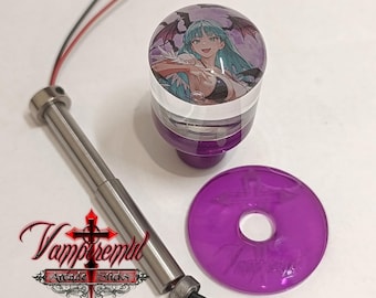 May include: A purple arcade joystick ball top featuring an anime character with teal hair and wings, a metallic shaft with attached wiring, and a translucent purple circular dust washer with an embossed cross and the text Vampremil Arcade Sticks.