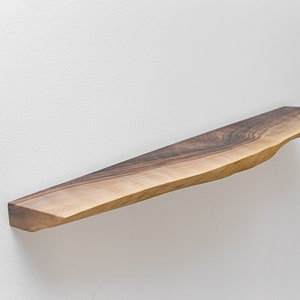 May include: A long, narrow wooden shelf with a natural, live edge finish. The shelf is made of dark brown wood with a light brown grain pattern. It is mounted to a white wall.