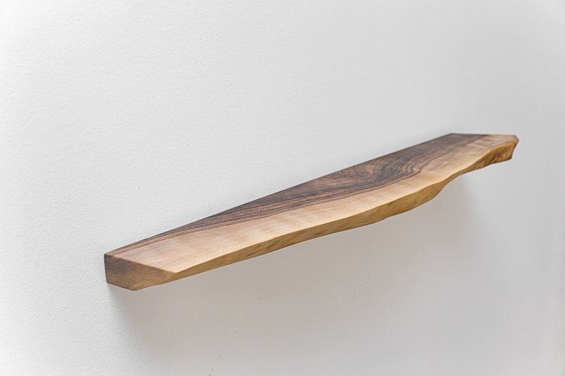 May include: A long, narrow wooden shelf with a natural, live edge finish. The shelf is made of dark brown wood with a light brown grain pattern. It is mounted to a white wall.