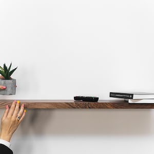 May include: A dark brown wooden shelf with a small potted succulent plant, a remote control, and two books on top. The shelf is mounted on a white wall.