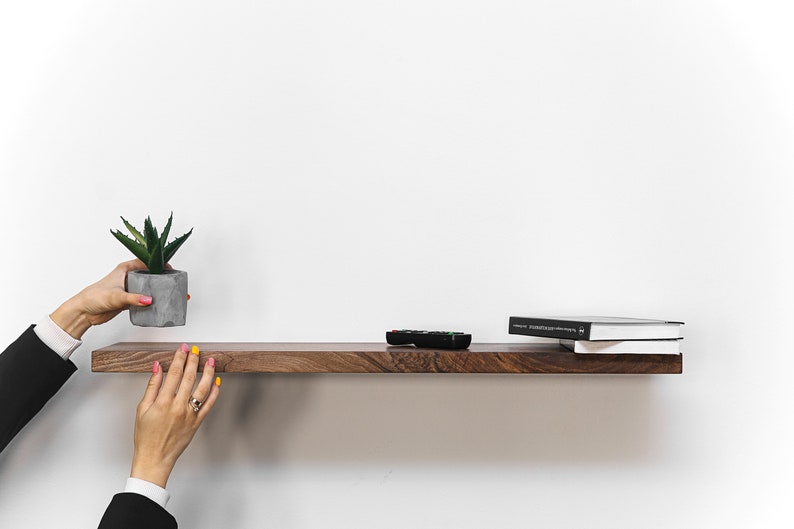 May include: A dark brown wooden shelf with a small potted succulent plant, a remote control, and two books on top. The shelf is mounted on a white wall.