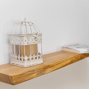 May include: A white birdcage with intricate details sits on a wooden shelf with a book on top. The shelf is mounted on a white wall.